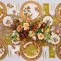 A top-down view of the Acorn and Oak Table Setting by Sophistiplate, featuring a fall-themed floral centerpiece, Thanksgiving tableware, woven placemats, gold flatware, and leaf-motif napkins in green, orange, and brown.