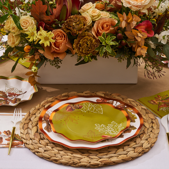The Acorn and Oak Table Setting by Sophistiplate features woven placemats, Thanksgiving tableware with autumn leaves, gold utensils, and a floral centerpiece with orange, yellow, and cream blooms for a festive fall tablescape.