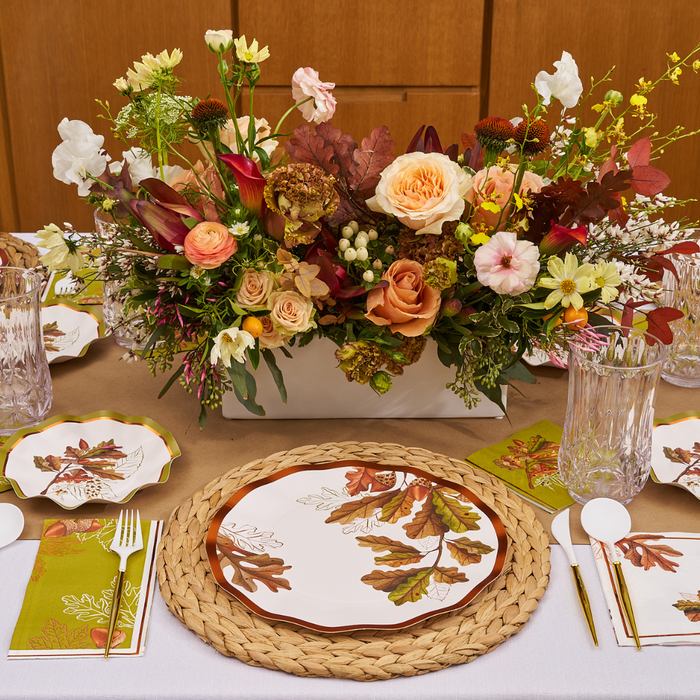 The Acorn and Oak Table Setting by Sophistiplate creates a stunning fall tablescape with a floral centerpiece, autumn leaf plates, woven placemats, gold flatware, green leaf napkins, clear glasses, and a tan tablecloth.