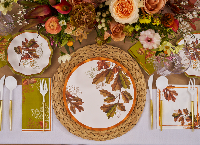 A fall tablescape shines with the Sophistiplate Acorn and Oak Table Setting—plates and napkins featuring leaf motifs, gold utensils, and clear glasses on a woven placemat over a tan tablecloth.