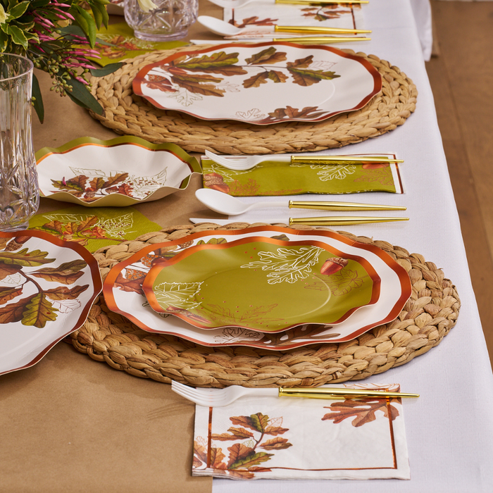 The Acorn and Oak Table Setting by Sophistiplate features disposable plates, napkins, and bowls with autumn leaves, green napkins, gold cutlery, and woven placemats arranged on a white tablecloth for a festive fall tablescape.