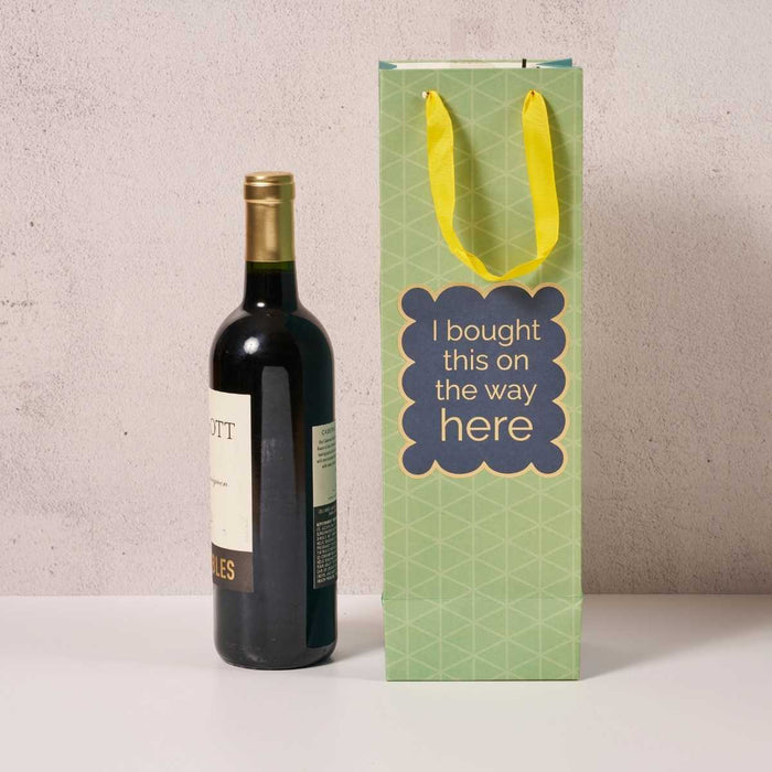 A bottle of wine sits beside the Drinks On Me “Bought This- Wine Bag,” a green, reusable bag with yellow handles and a blue label reading, “I bought this on the way here,” shown against a plain light-colored background.
