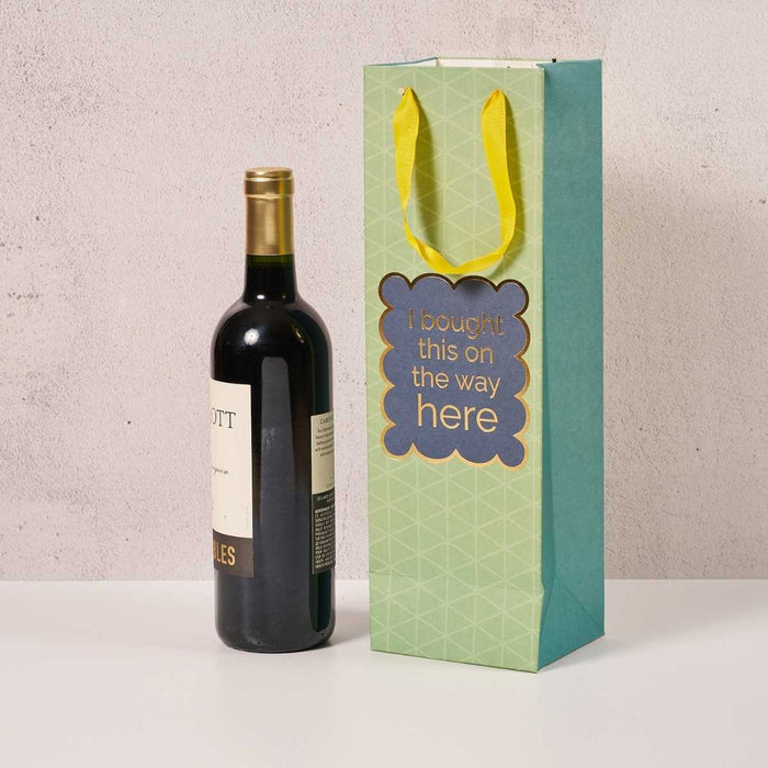 A bottle of red wine sits beside the Drinks On Me Bought This- Wine Bag, featuring yellow handles and "I bought this on the way here" on the front—ideal as a hostess gift.