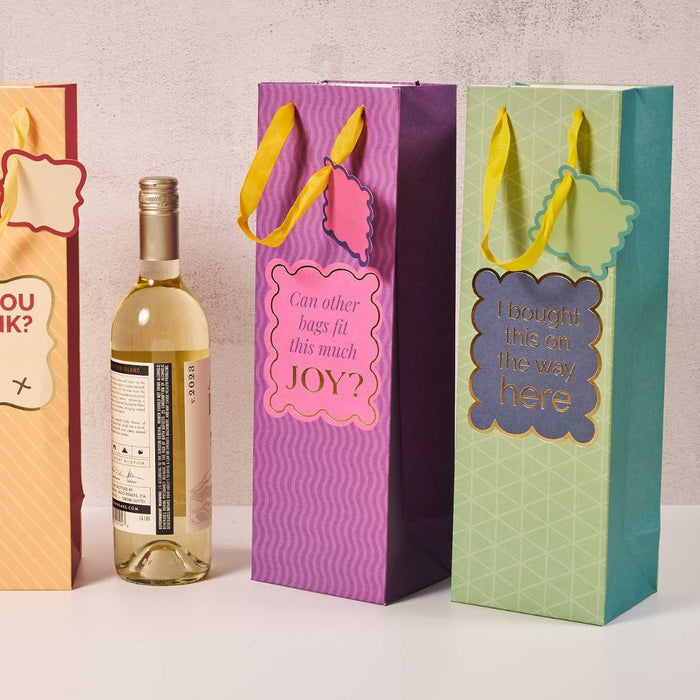 Three colorful Drinks On Me hostess gifts with humorous tags stand next to a bottle of white wine, including the Bought This- Wine Bag with yellow ribbon handles and the label: I bought this on the way here.