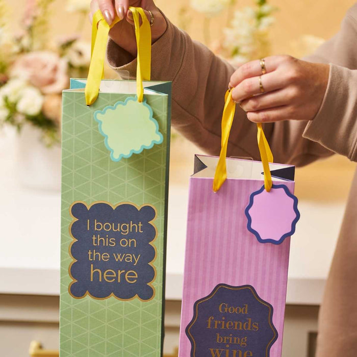 Two hands hold two Drinks On Me wine bags with yellow ribbon handles—one green bag says “I bought this on the way here,” and one purple bag says “Good friends bring wine.” Both feature attached gift tags, making them perfect hostess gifts.