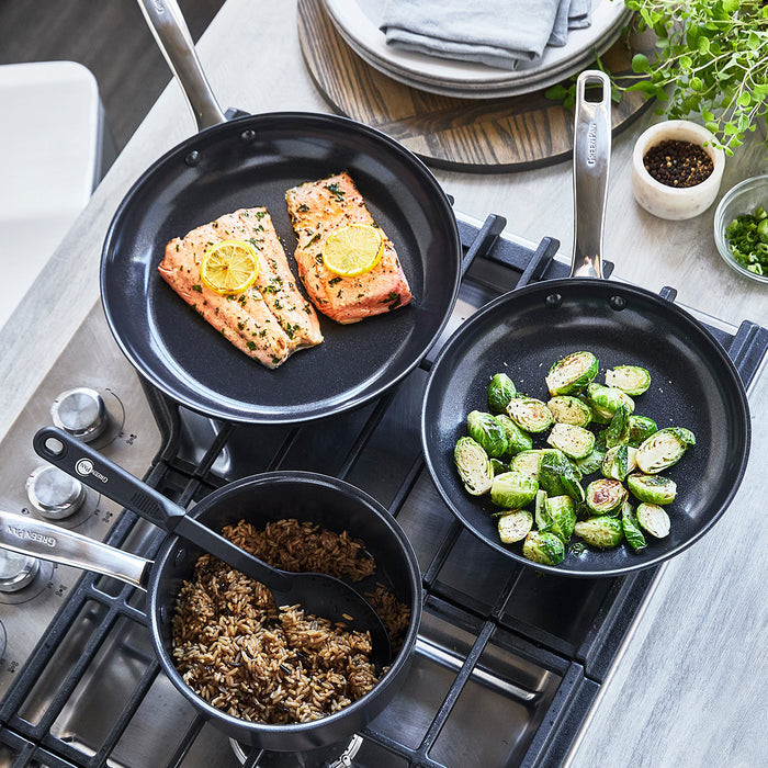 Black ceramic 5-piece cookware set with three pans on a stove, each holding a cooked dish.