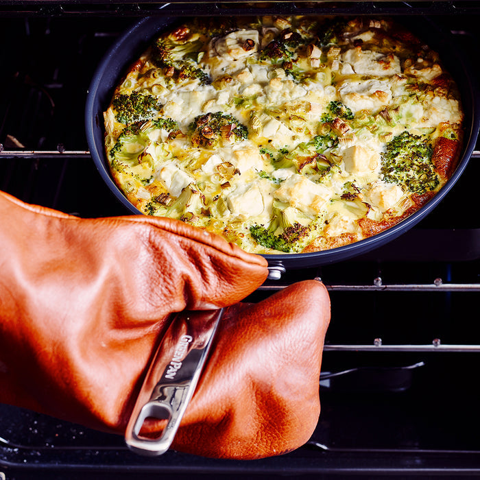 A hand with an oven mitt pulls a broccoli and cheese frittata from a black ceramic nonstick pan.