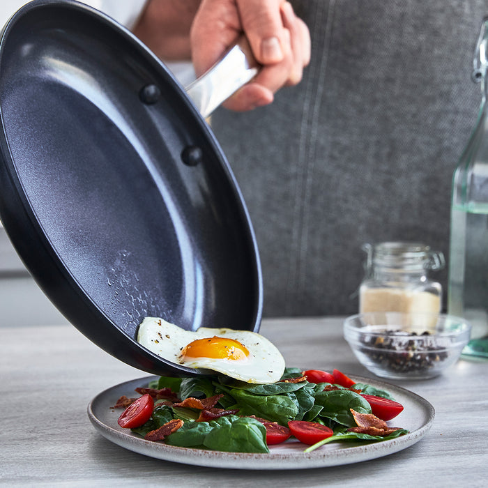 Fried egg in a black ceramic nonstick pan, served over spinach and tomatoes on a kitchen counter.