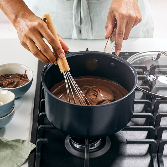 Melted chocolate being whisked in a black ceramic saucepan with mixing bowls nearby on the counter.