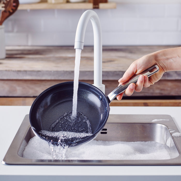 Hand rinsing a black ceramic nonstick pan under running water above a soapy kitchen sink.