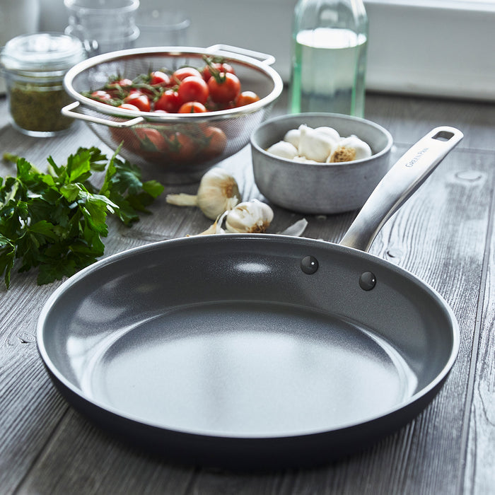 The Chatham Ceramic Nonstick 10" Frypan with a stainless steel handle rests on a wooden table beside parsley, garlic bulbs, cherry tomatoes in a colander, and a glass bottle, ready for food prep.