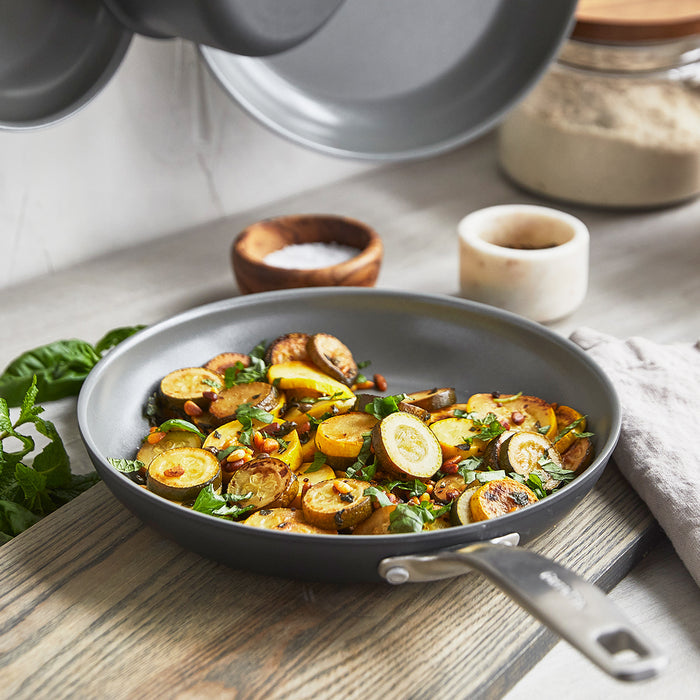 The Chatham Ceramic Nonstick 10" Frypan with a stainless steel handle holds sautéed yellow squash, herbs, and pine nuts, and sits on a kitchen counter beside a salt cellar, glass jar, and folded beige cloth.