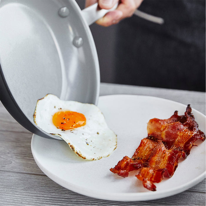 A hand slides a fried egg from the Chatham Ceramic Nonstick 10" Frypan onto a white plate beside several strips of cooked bacon on a wooden surface.