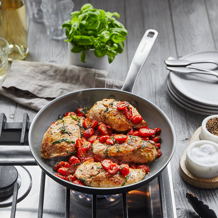 The Chatham Ceramic Nonstick 10" Frypan with cooked chicken breasts and cherry tomatoes sits on the stovetop, its stainless steel handle gleaming amid fresh basil, stacked plates, cutlery, salt, pepper, and glasses on a wooden counter.