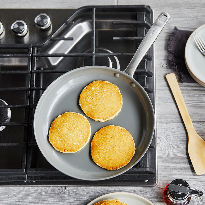 The Chatham Ceramic Nonstick 10" Frypan with a stainless steel handle cooks three golden pancakes on a stovetop, shown beside a wooden spatula, stacked plates with utensils, and a syrup bottle on a light wood surface.