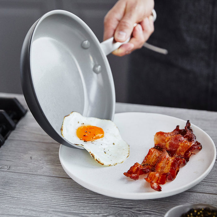 A hand slides a sunny-side-up egg from a white ceramic frying pan onto a plate with bacon.