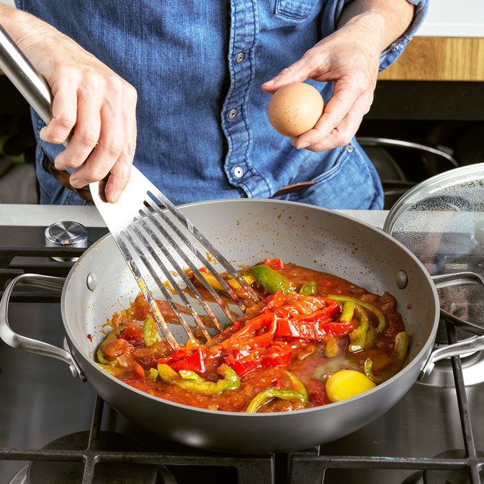 Hand holding an egg over a Chatham ceramic nonstick 11-inch pan with tomato sauce, peppers, and a spatula inside.