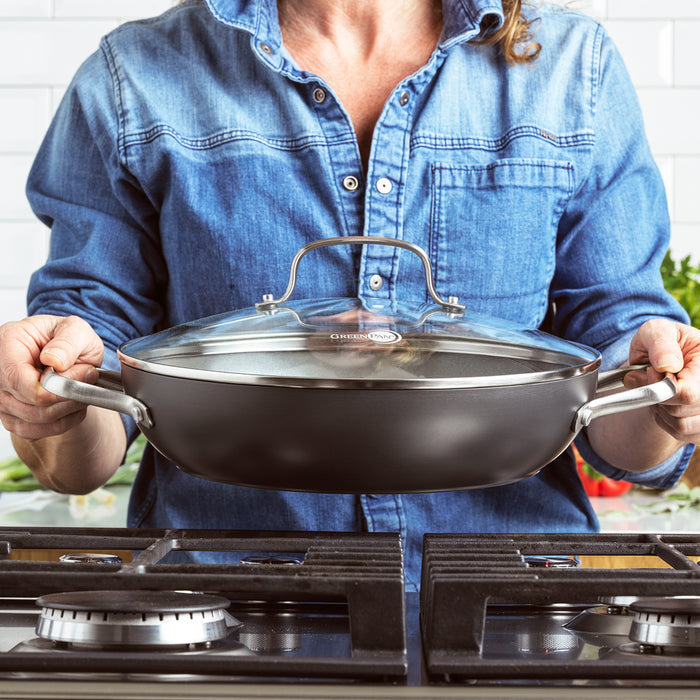 Person in a denim shirt holding a ceramic nonstick 11 pan with lid over a stove in a kitchen.