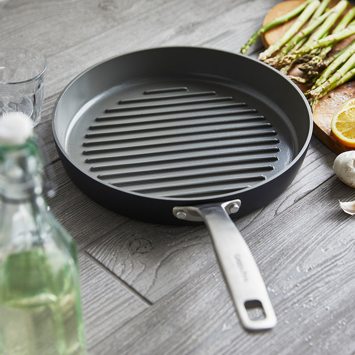 Ceramic nonstick grill pan on wood surface with asparagus, lemon, garlic cloves, and a glass bottle nearby.