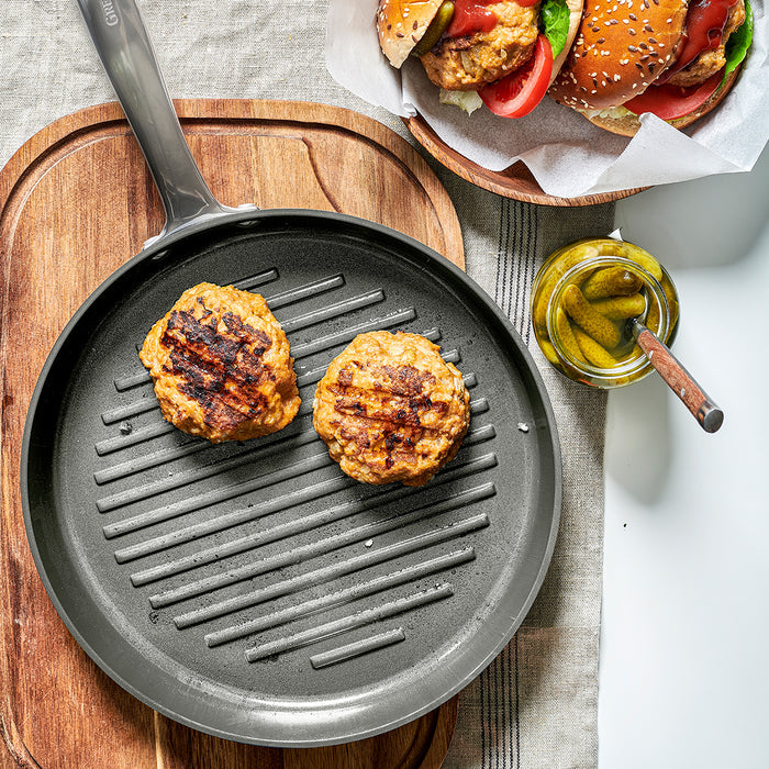 Two burger patties cooking in an 11-inch Chatham ceramic nonstick grill pan, with pickles and buns on the side.