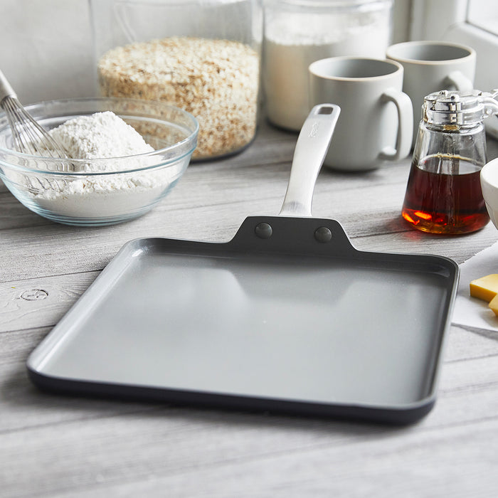 Square ceramic nonstick griddle on a kitchen counter with flour, oats, and syrup placed nearby.