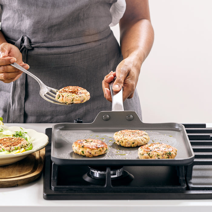 Person in gray apron flips patties on Chatham ceramic nonstick square griddle, with cooked patties beside it.