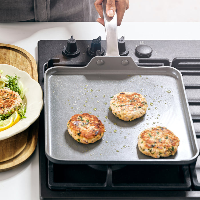 Three burger patties cooking on a square grey ceramic griddle with a hand nearby and a bowl of salad in the background.