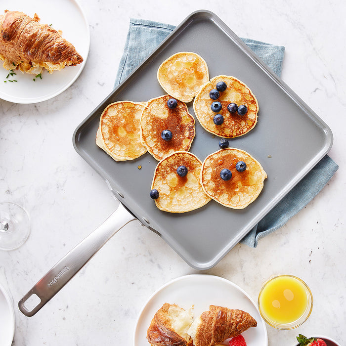 Square ceramic nonstick griddle with several small pancakes, some topped with blueberries and syrup, on a table.