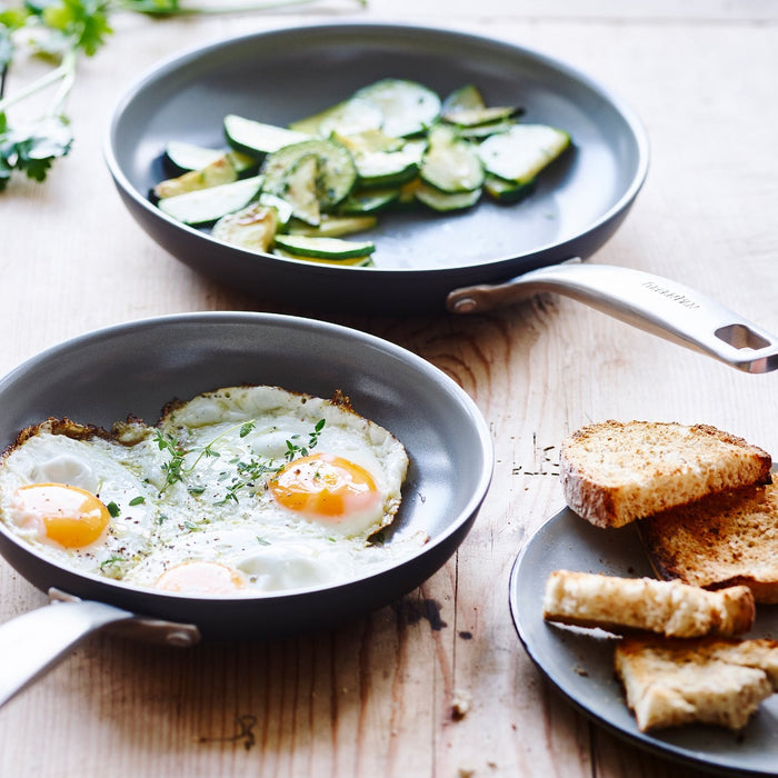 Two ceramic nonstick frypans, 8 and 10, cooking eggs and zucchini, next to toast on a wooden table.