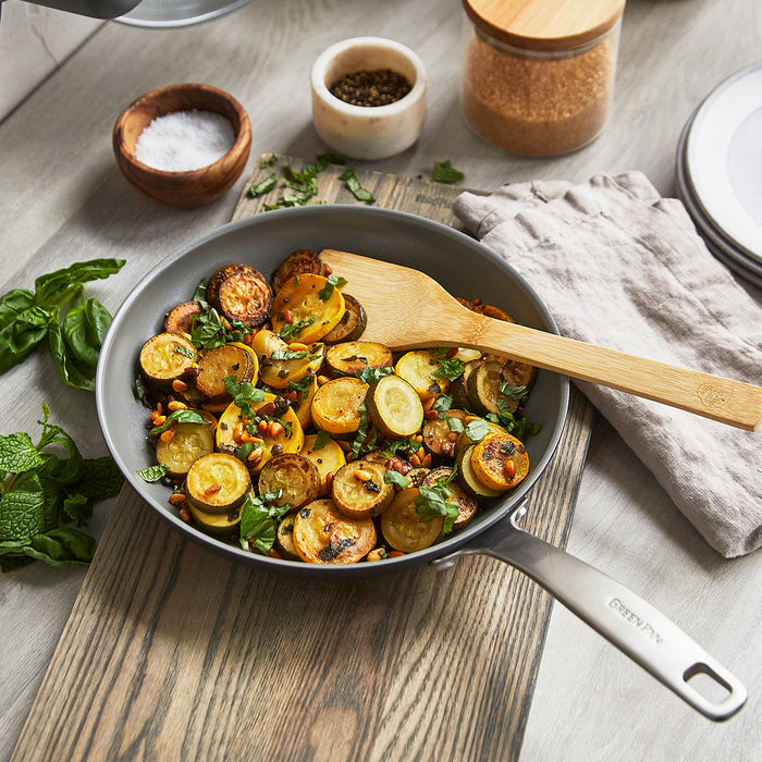 Green pan with sautéed zucchini and herbs, next to a wooden spatula and two small spice bowls on a countertop.