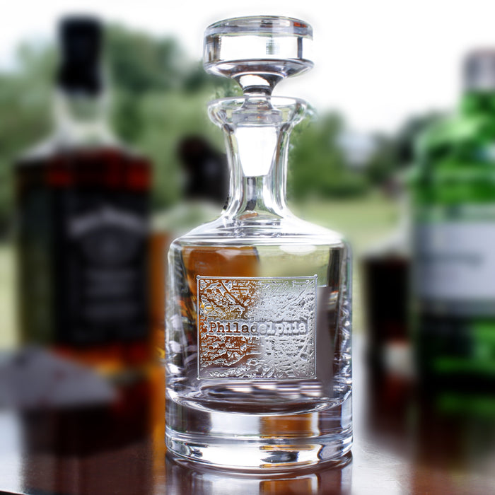 A glass spirits decanter deep-carved with a Philadelphia street-map panel and the city name, with whiskey bottles blurred behind.
