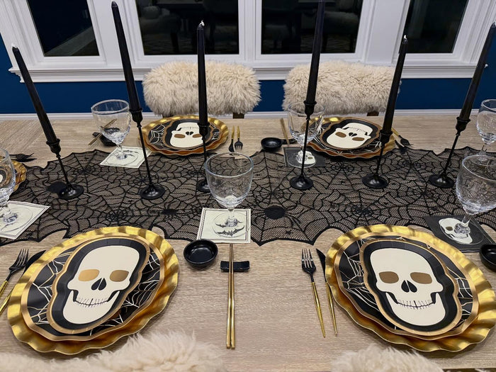 A Halloween-themed tablescape features Sophistiplate’s Classic Spooky Skull Salad Plates (8pk), gold utensils, black candles, party plates, a spiderweb tablecloth, and clear glasses—all perfectly arranged and ready for a delightfully eerie dinner party.