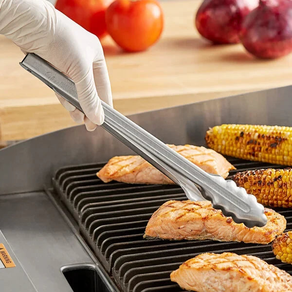 Chef grilling chicken and corn using tongs on a barbecue with tomatoes in background.