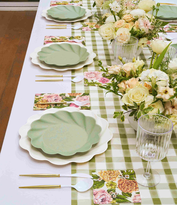 The Garden Rose Table Setting by Sophistiplate features pastel green scalloped plates, gold cutlery, ribbed glasses, and floral napkins on a green gingham tablecloth, with white and pale pink floral centerpieces.