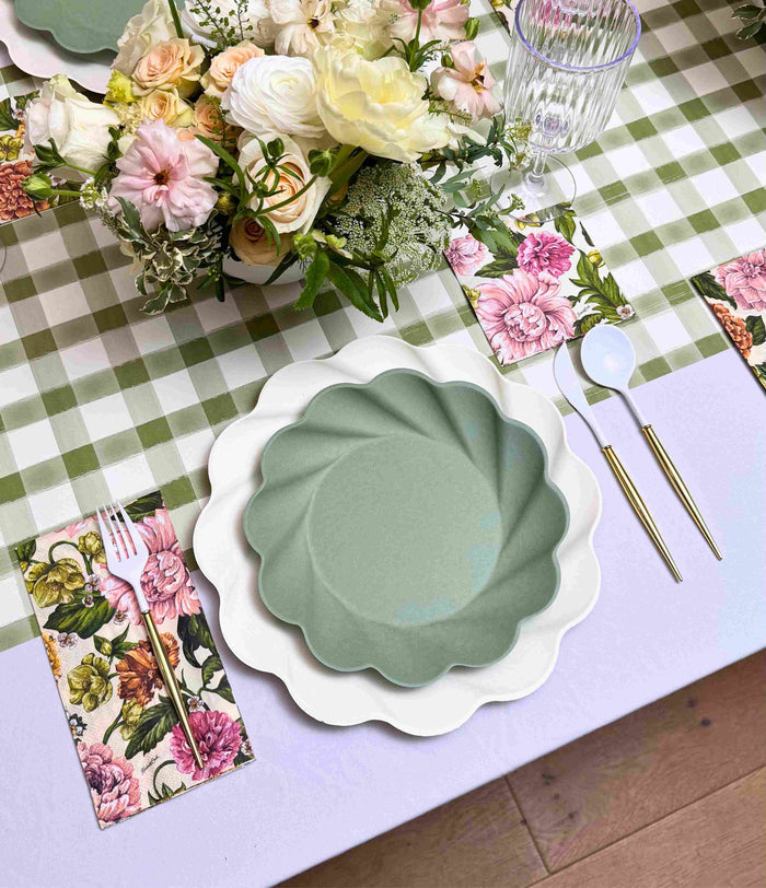 The Garden Rose Table Setting by Sophistiplate features white and green scalloped designer plates, gold cutlery, a clear glass, floral napkins, and pastel flowers on a green gingham tablecloth.