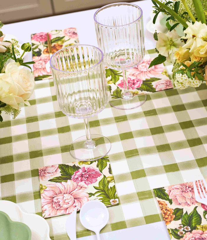 The Garden Rose Table Setting by Sophistiplate features green and white checkered placemats, pink floral napkins, clear ribbed wine glasses, white utensils, and decorative disposable plates with white and pale yellow floral arrangements.