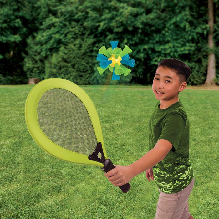 A smiling boy in a green shirt plays GO! Play Bashminton by GO! Play, swinging an oversized paddle at a colorful foam ball while standing on grass in a park with trees, enjoying outdoor fun.