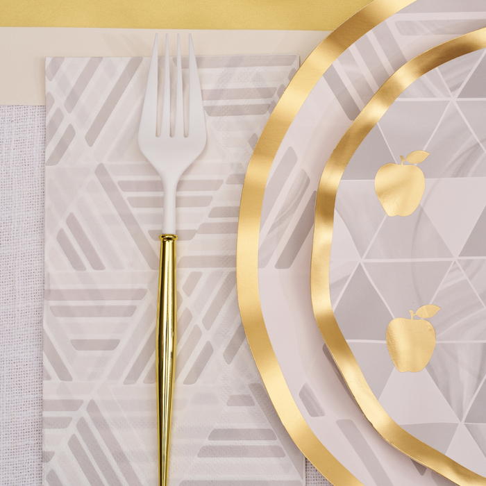 A white plastic fork with a gold handle rests on a geometric cocktail napkin beside Sophistiplate Golden Apple Guest Towels, featuring plates adorned with gold apples and gold foil trim, all arranged in an elegant table setting of neutral and gold hues.