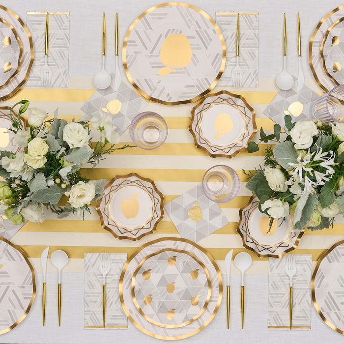 Sophistiplate Golden Apple Cocktail Napkins (26pk) add a touch of glamour to an elegant table setting featuring a gold-striped tablecloth, geometric-patterned plates, and gold utensils. The look is completed with crisp white napkins and floral centerpieces of white and green blooms, all artfully arranged for a sophisticated, symmetrical presentation.