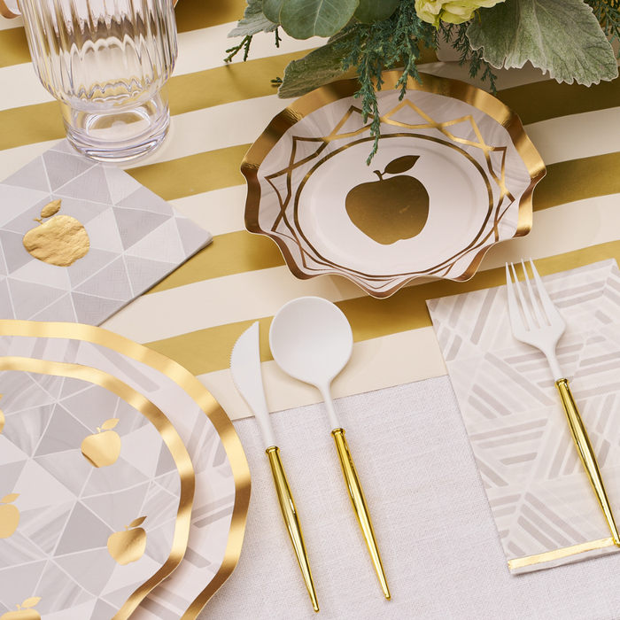 A Golden Apple Table Setting features Sophistiplate’s Golden Apple Guest Towels, adding a sophisticated touch with their elegant gold foil trim and apple motif. These guest towels are beautifully complemented by gold-accented cutlery, geometric cocktail napkins for a modern flair, a glass, and a floral centerpiece—all artfully arranged on a striped table runner.