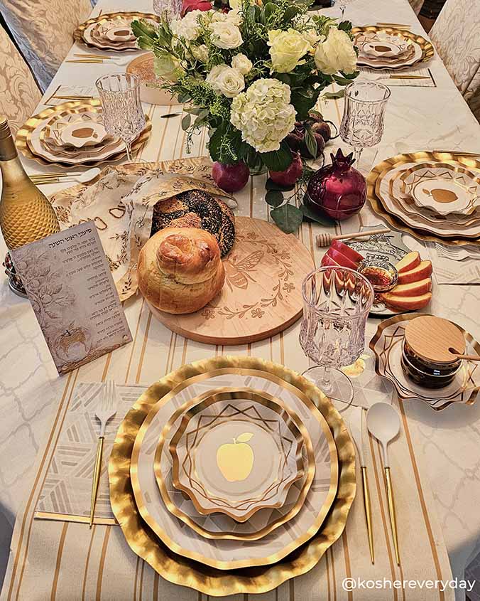 A festive table is set with gold and white plates featuring gold foil trim, glasses, a wine bottle, round challah, apples, honey, a pomegranate, and a floral centerpiece; a prayer card is also included. Sophistiplate’s Golden Apple Guest Towels (20pk) add a modern touch to the celebratory and elegant Golden Apple Table Setting.