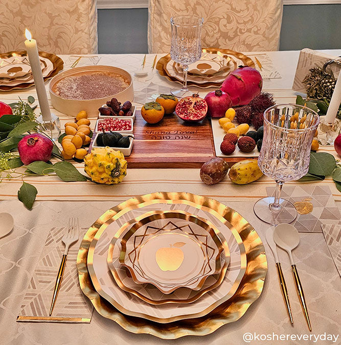 A festive holiday table is set with gold-rimmed plates, crystal glasses, gold cutlery, candles, pomegranates, and assorted fruits arranged atop a cream tablecloth. Adding an extra touch of elegance to this Golden Apple Table Setting are the Sophistiplate Golden Apple Cocktail Napkins (26-pack).
