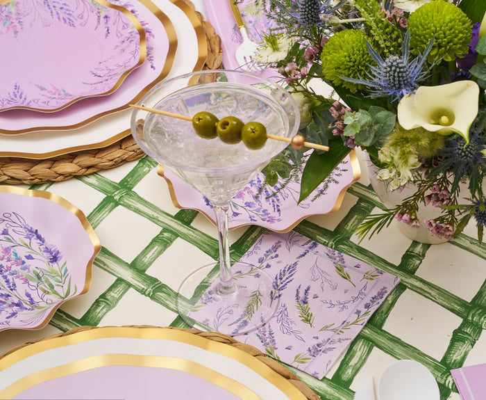 A martini with three green olives rests on a floral-themed table set with lavender plates, gold-edged chargers, a wicker placemat, floral napkin, and a bouquet of purple, green, and white flowers atop a Hester & Cook Green Lattice Runner (20x25").