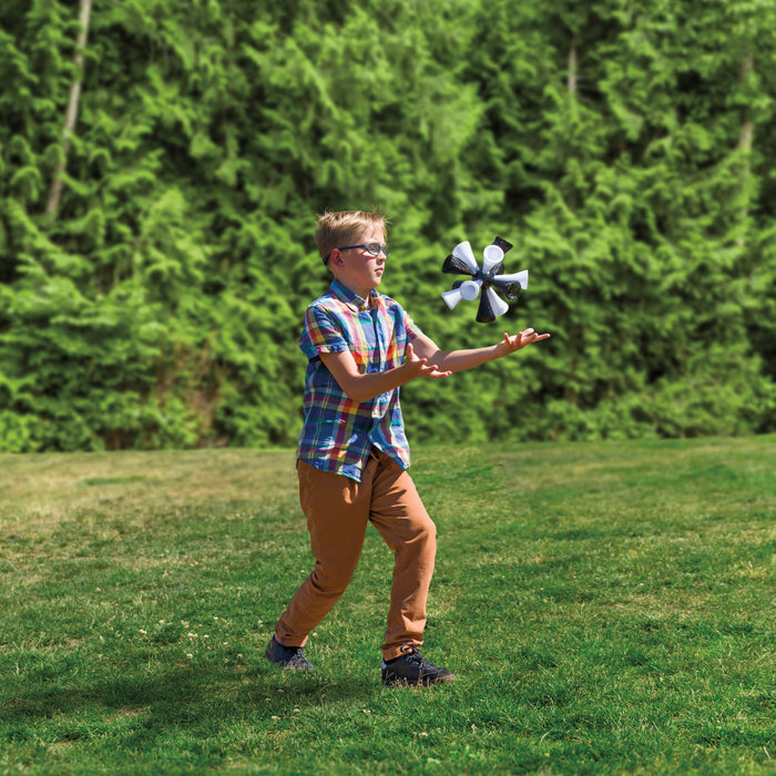 A boy in glasses, a plaid shirt, and tan pants plays outdoors on grass, reaching up to catch the GO! Play Jumbo Bash Birdie in the backyard with tall green trees behind him.