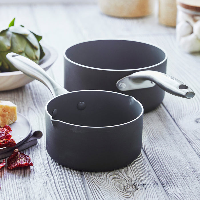 Two gray ceramic saucepans with silver handles on a wooden countertop, shown with assorted cooking ingredients.