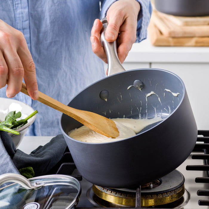 White sauce being stirred with a wooden spoon in a cream-colored ceramic saucepan on a gas stove.