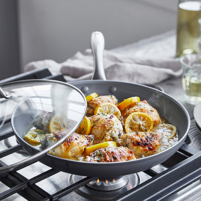 Chicken thighs with lemon cook in a ceramic nonstick 12-inch frypan with lid; wine glass sits in the background.