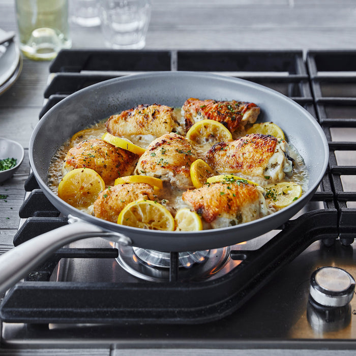 Chicken thighs with lemon slices in sauce and herbs cooking in a ceramic nonstick 12-inch frypan with lid.
