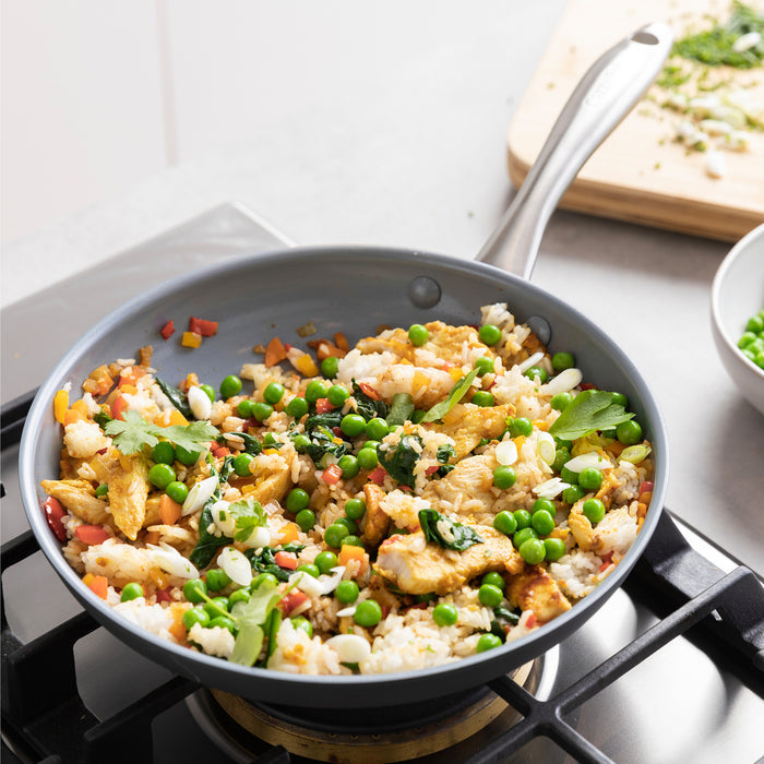 Gray ceramic nonstick frypan with glass lid on stove, filled with fried rice, peas, carrots, eggs, and onions.
