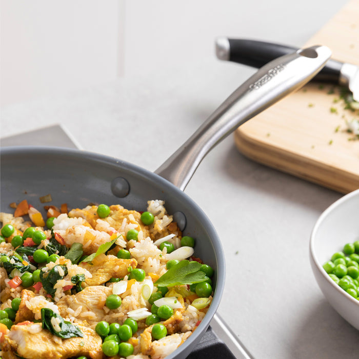 Fried rice with peas and vegetables in a green ceramic nonstick frypan with glass lid on a stovetop.